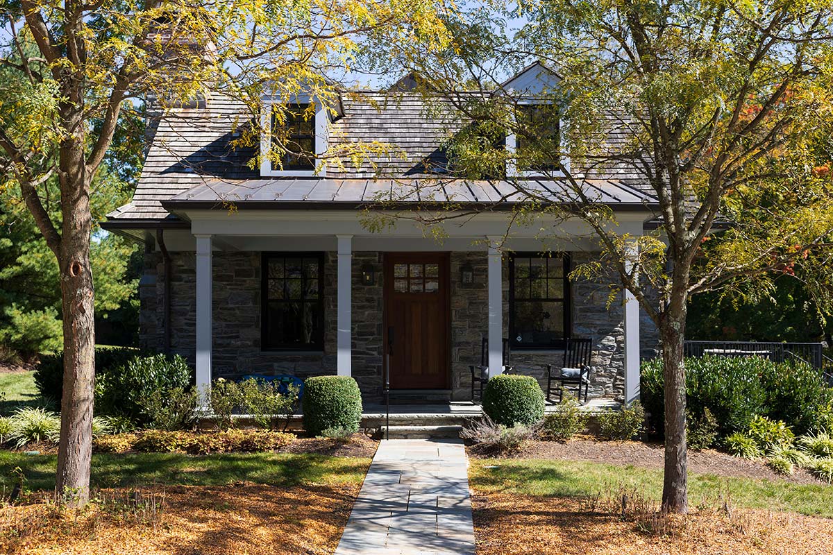 Cozy stone house with a gabled roof, wooden door, and porch with columns, surrounded by trees and a stone path in a lush garden.