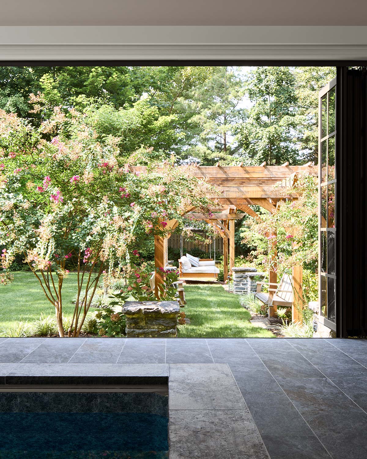 View from an indoor pool room with large open windows to a garden with a wooden pergola, swings, green lawn, and blooming shrubs.