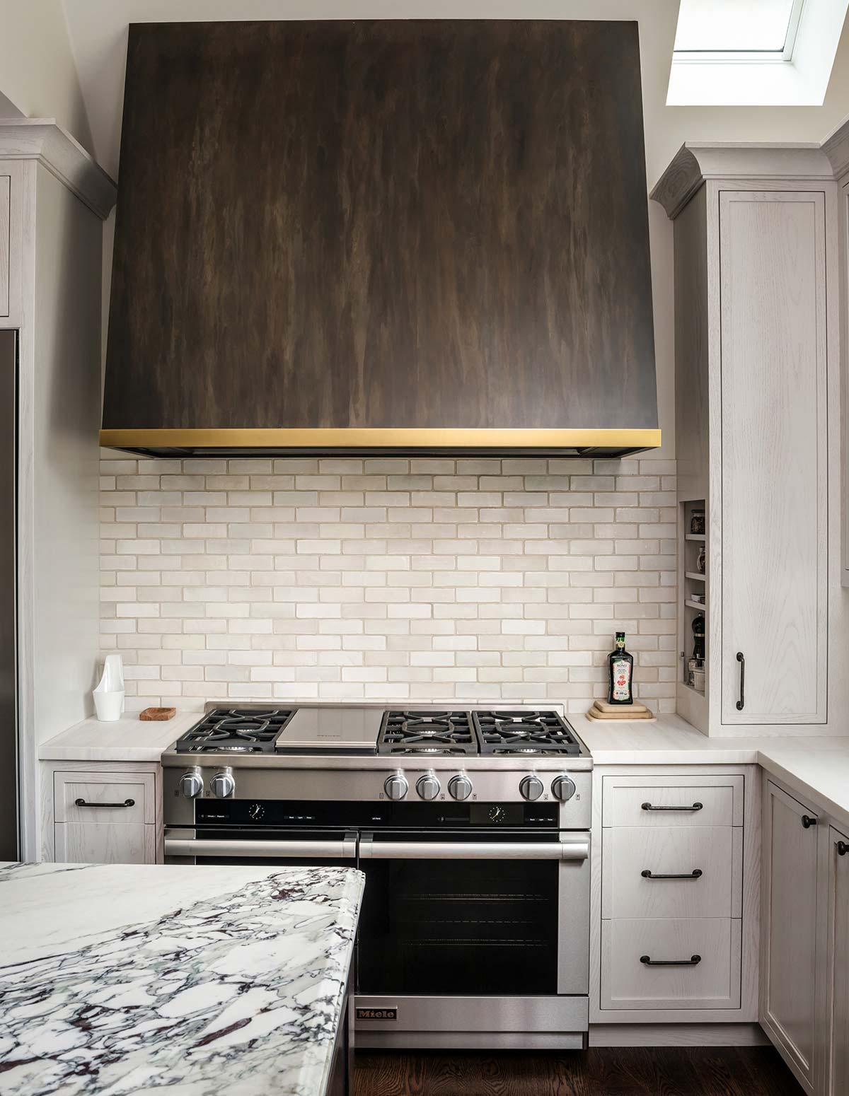 Modern kitchen with a dark wood hood, marble countertop, stainless steel gas stove, white cabinets, and a skylight above.