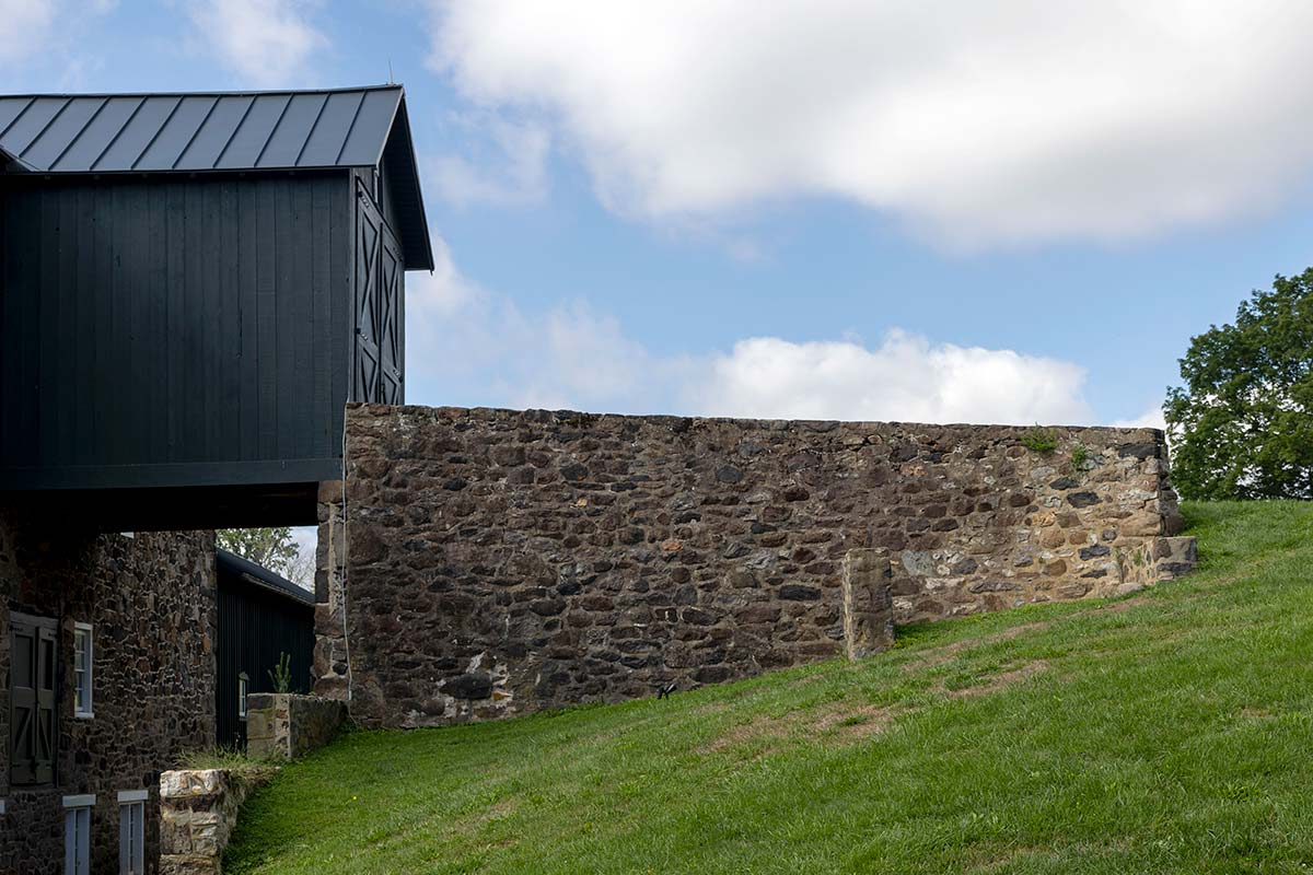 Black barn with metal roof next to a stone wall, on a grassy hill under a partly cloudy sky with trees in the background.