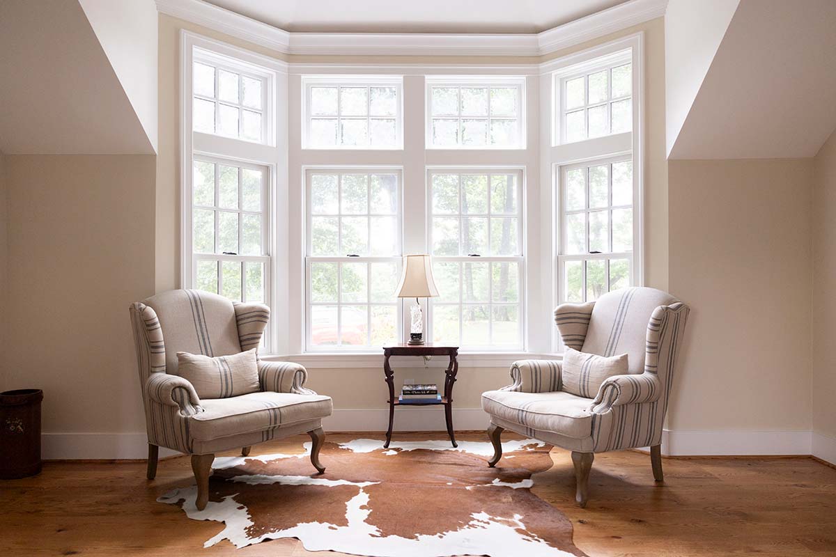 Cozy room with two striped armchairs, a small table with a lamp, and a cowhide rug, framed by large bay windows with natural light.