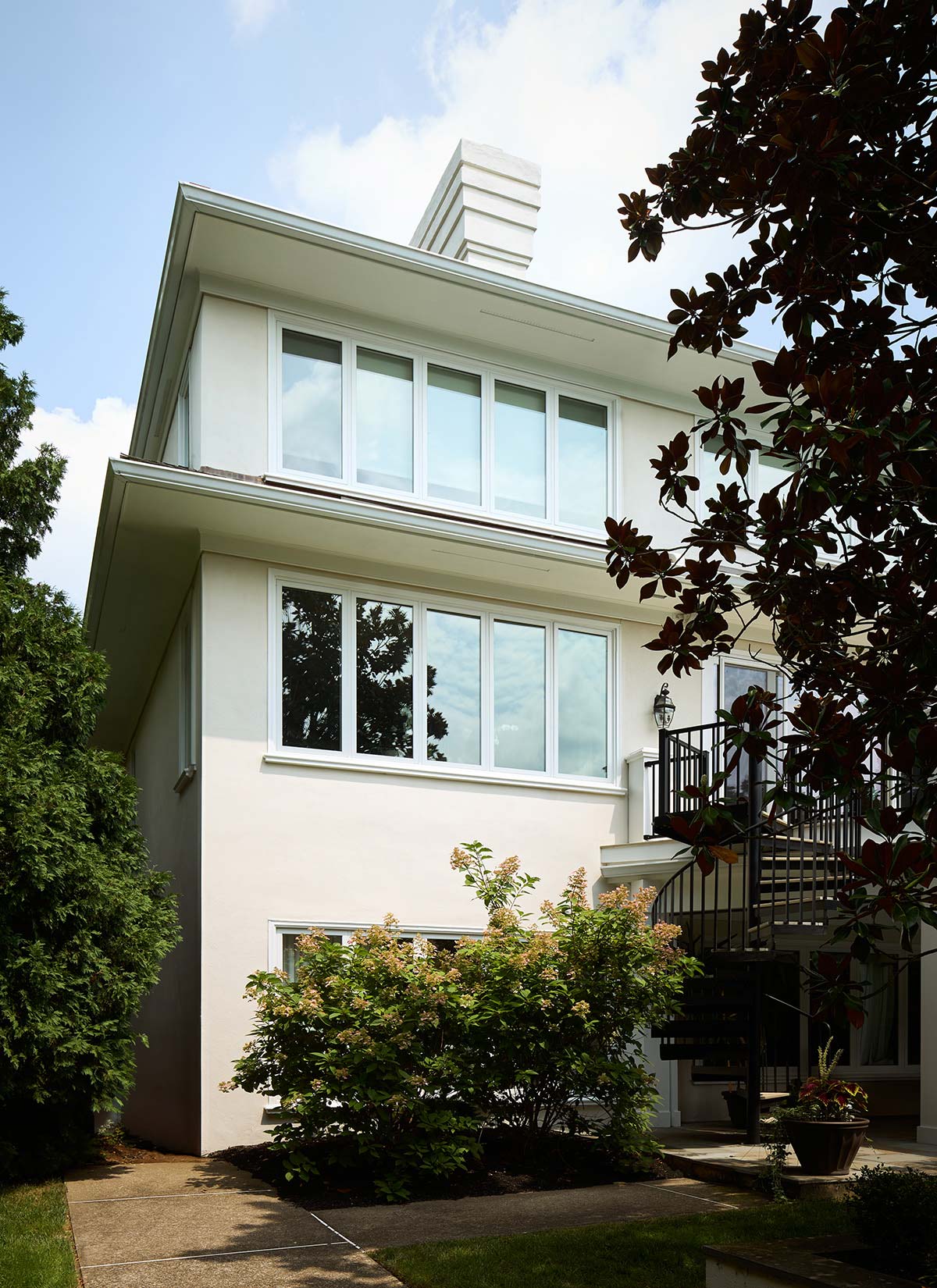 A three-story house with large windows, a spiral staircase, white exterior, and lush landscaping.