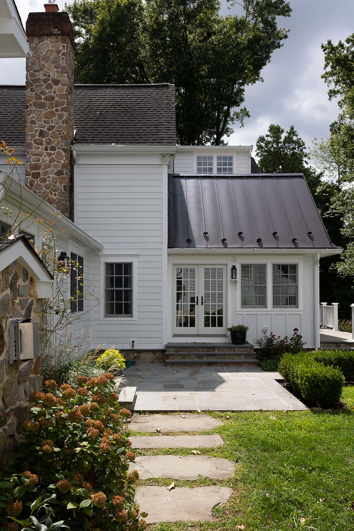 White house with stone chimney, metal roof extension, glass door, and a stone path through a lush garden with trees.