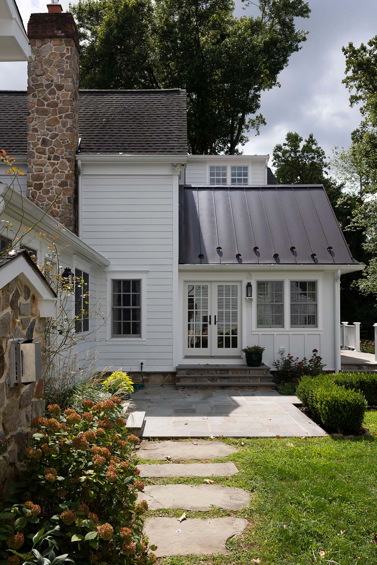 White house with stone chimney, metal roof extension, glass door, and a stone path through a lush garden with trees.