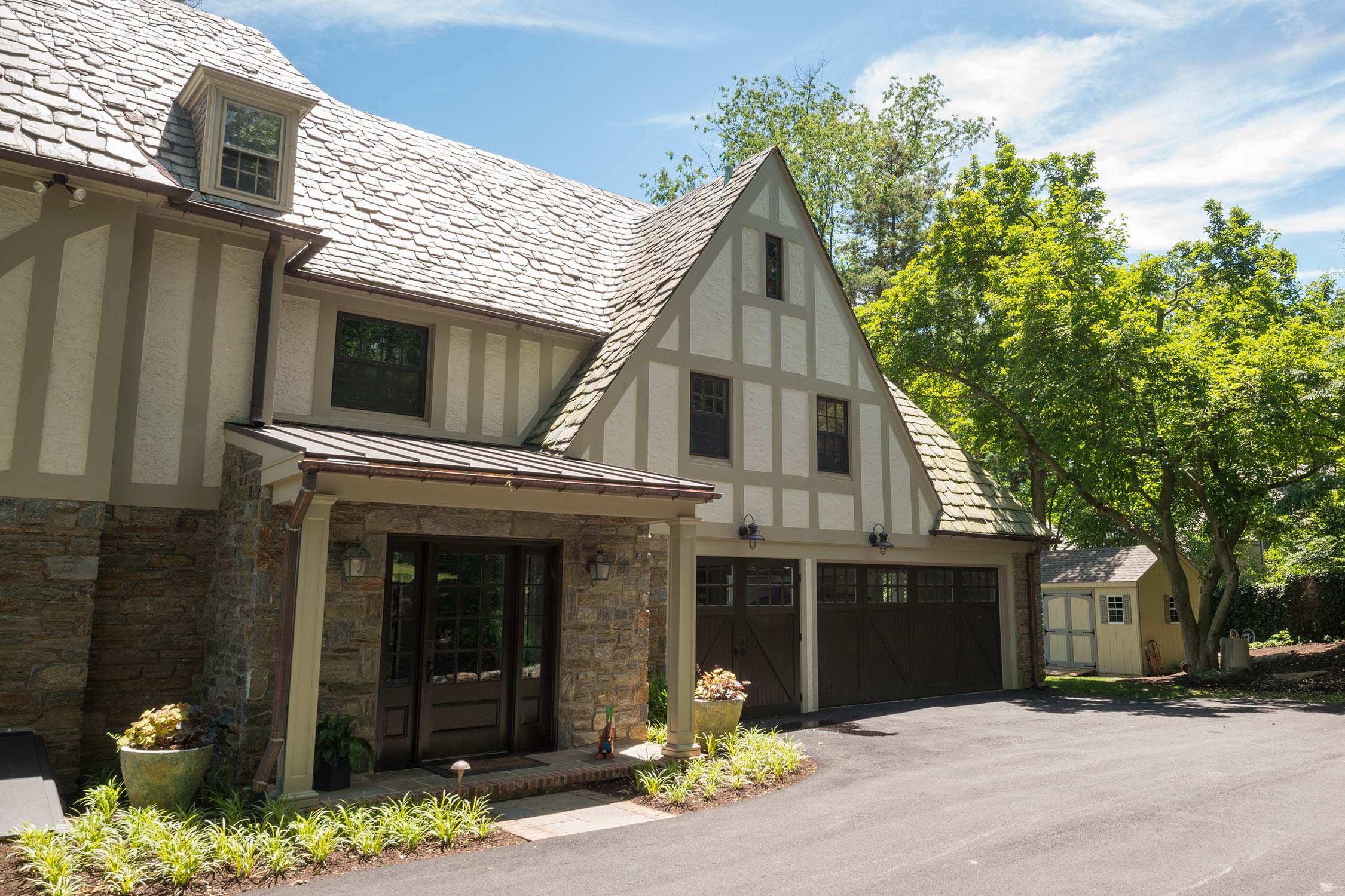Inviting stone Tudor home with slate roof, covered entry, double garage, and small shed, enhanced by manicured plants and blue skies.