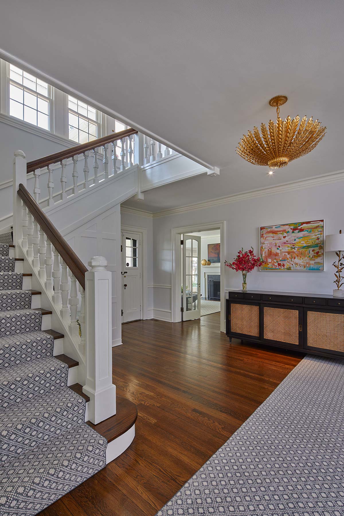 An elegant foyer with a grand staircase, hardwood floors, a gold chandelier, a wicker console with flowers, and colorful abstract art.