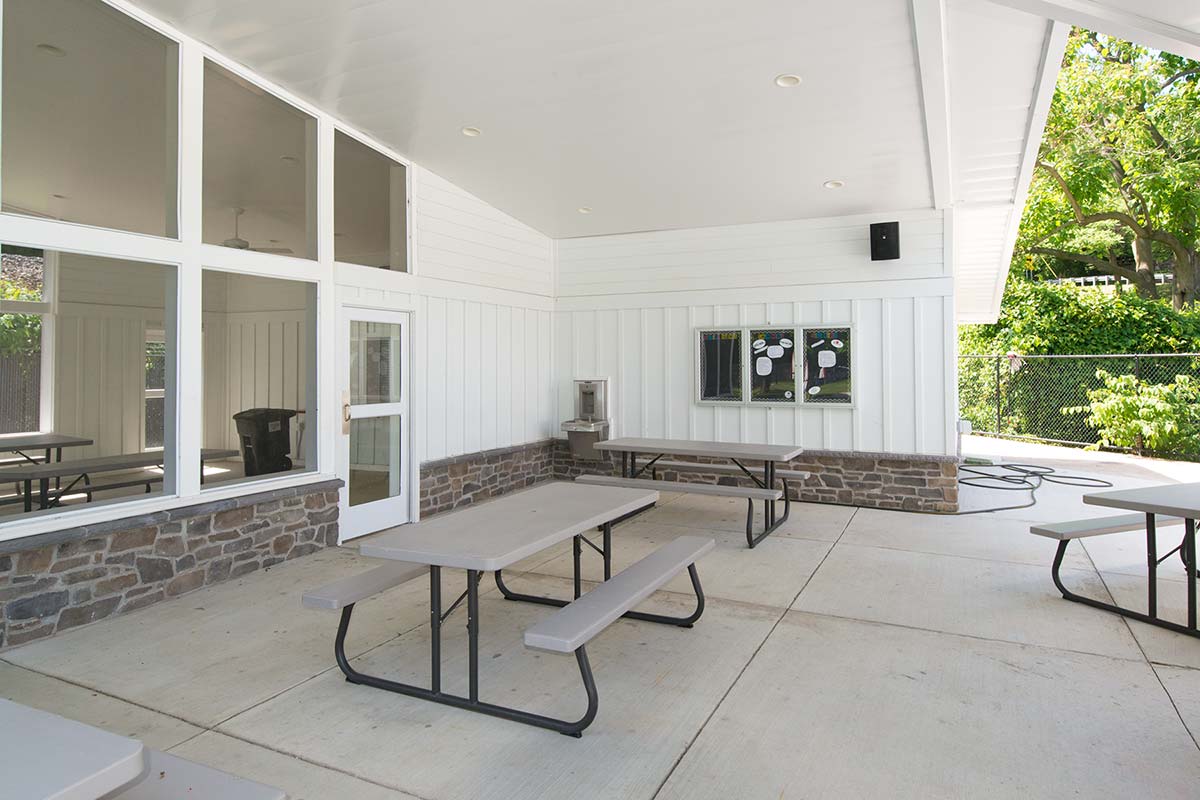 Covered outdoor area with picnic tables, white building, and bulletin board.