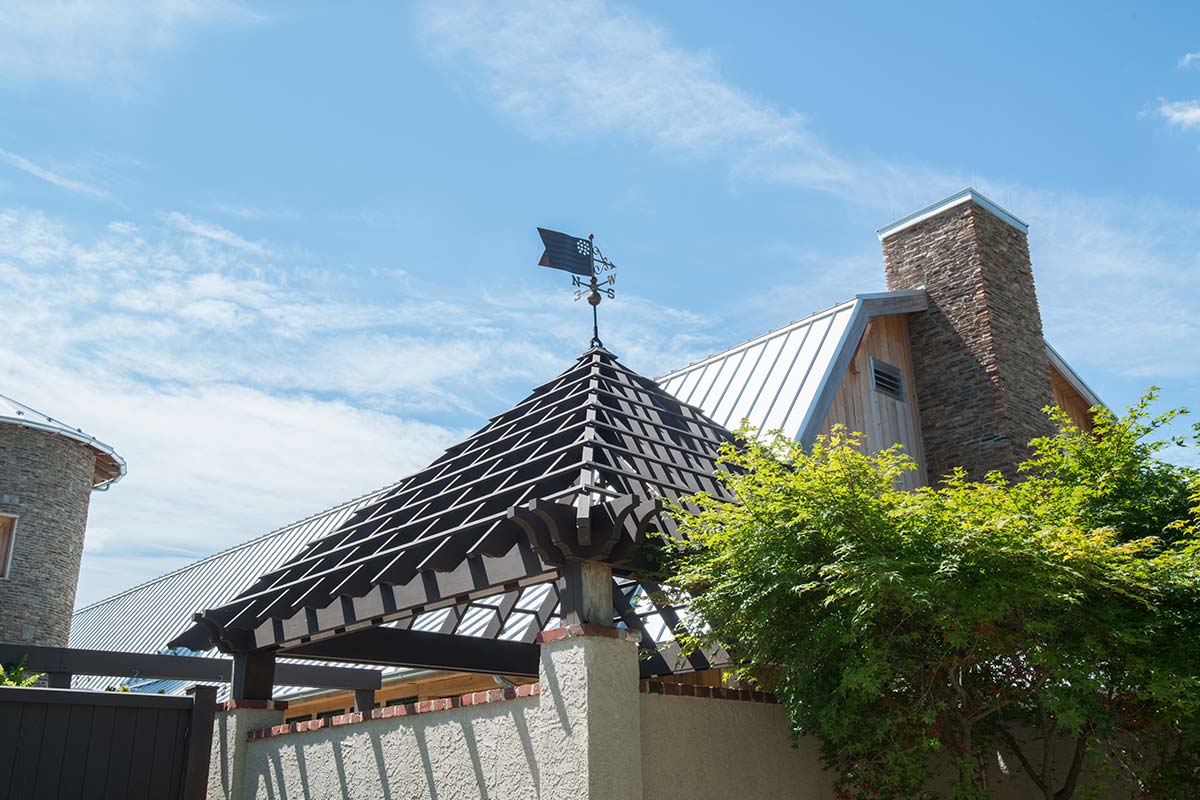 A pergola with a weather vane atop a stone chimney and metal-roofed building, surrounded by greenery under a blue sky.