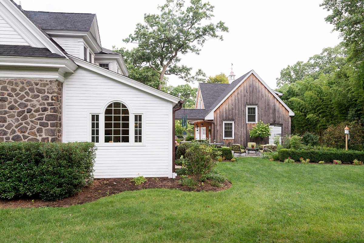 A white and stone house with a wooden barn, connected by a covered walkway, set in a lush green lawn with shrubs.