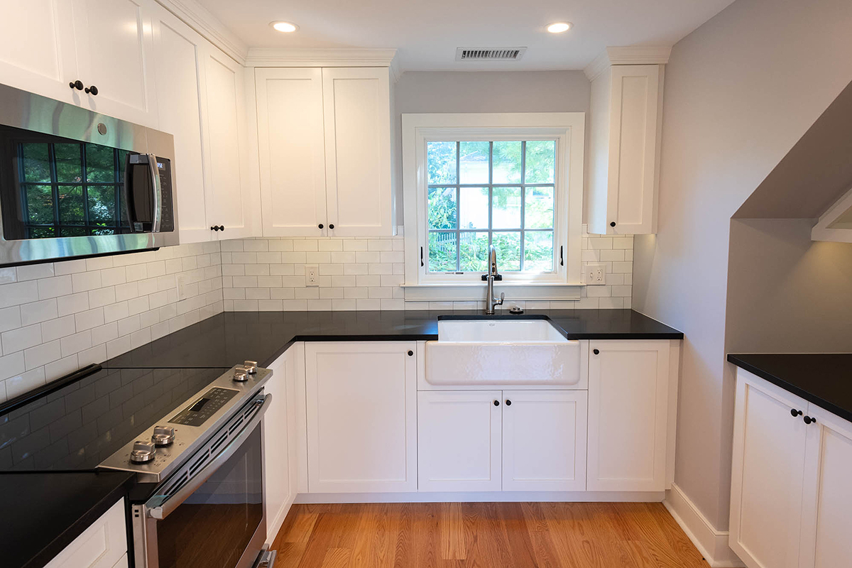 Compact cottage kitchen with white cabinets, black countertops, stainless appliances, farmhouse sink, subway tile, and window to greenery.