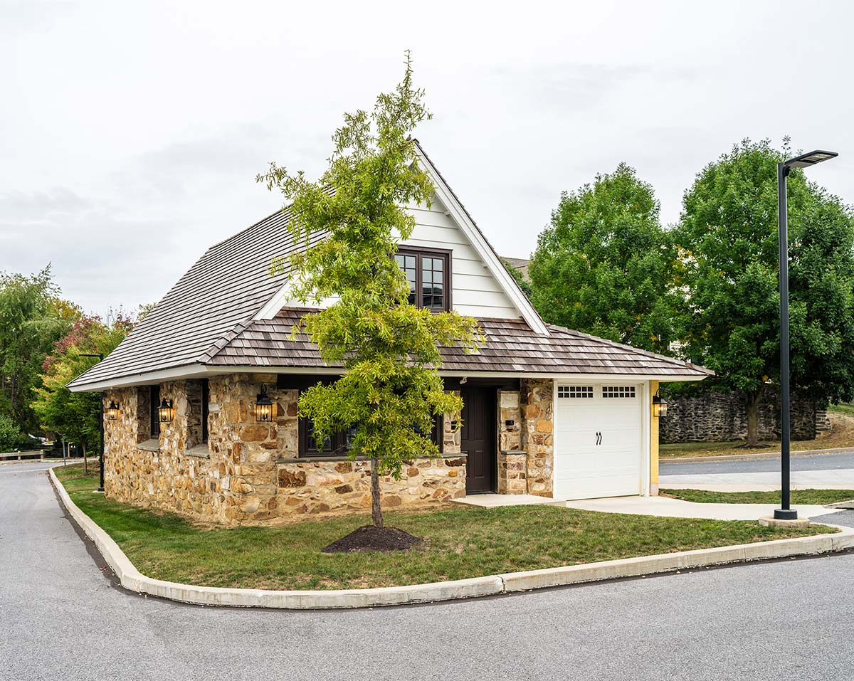 Stone and white garage with shingle roof, garage door, and tree in front, on a street with a lamppost.
