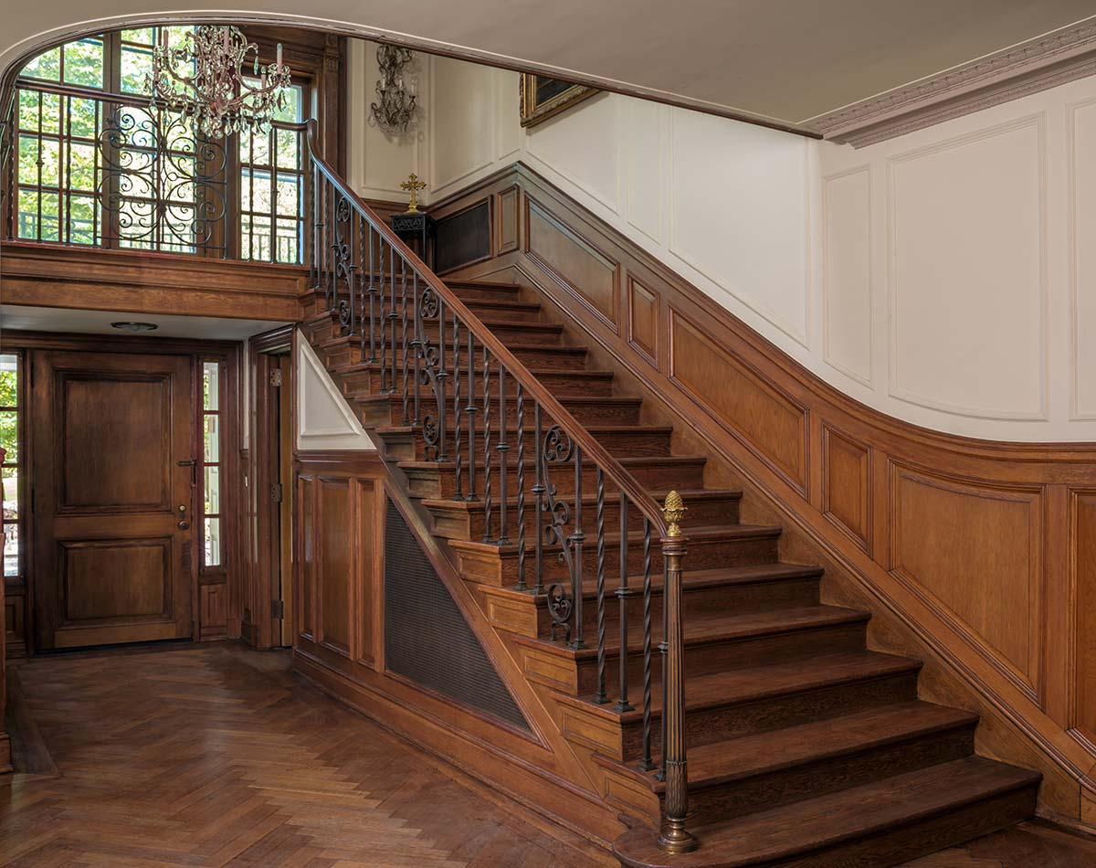 Grand foyer with wooden staircase, wrought iron railing, and chandelier. Features wood-paneled walls, a large arched window, and herringbone floor.