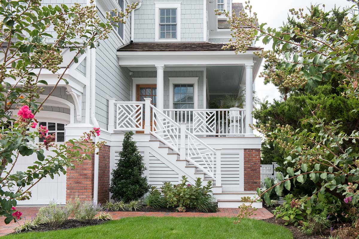 Beach house with gray shingles, white porch, and brick accents. Steps lead to the entrance, surrounded by greenery and pink flowers.