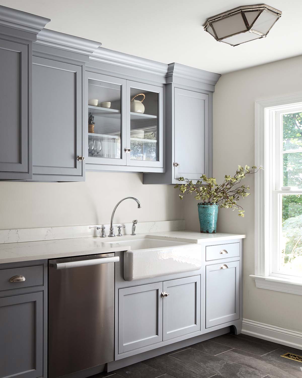Gray kitchen with farmhouse sink, stainless dishwasher, blue vase with greenery, and window.