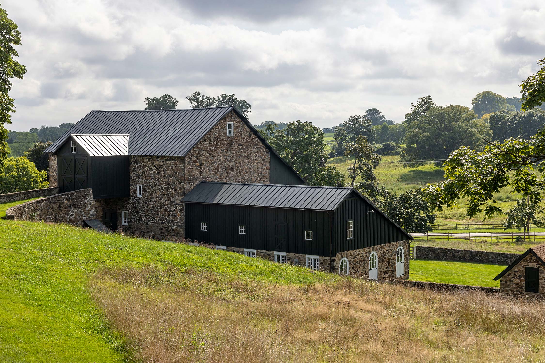 Stone and black metal barn with sloped roofs on a grassy hill, surrounded by trees and fields under a cloudy sky.