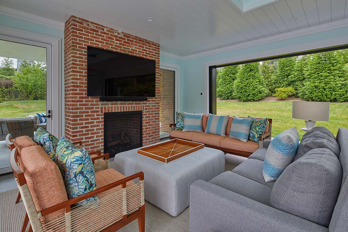 A cozy outdoor room with a brick fireplace, TV, gray sofa, orange chairs, and blue patterned pillows. Large windows show a green lawn outside.