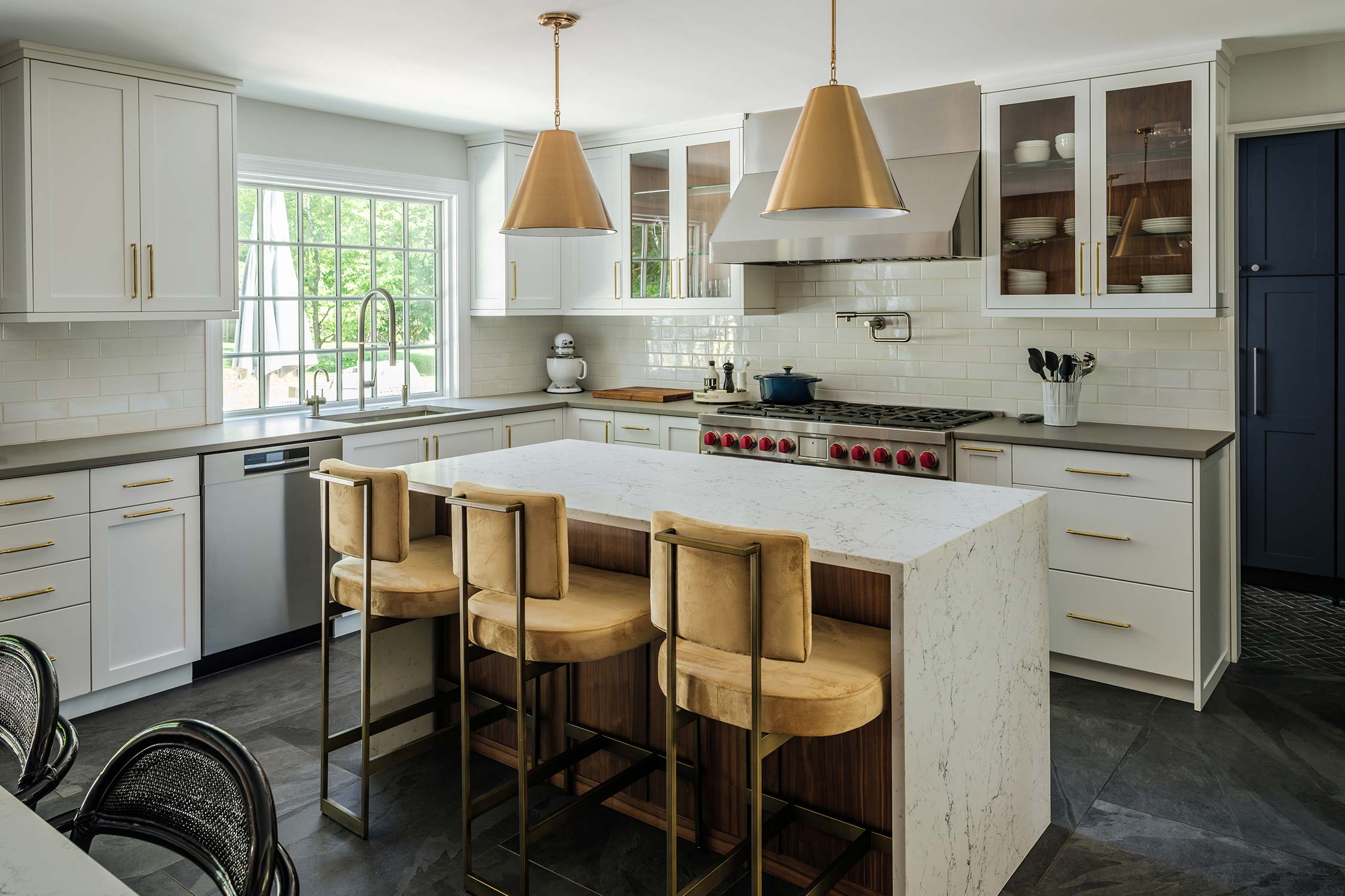 Modern kitchen with white cabinets, marble island, gold pendant lights, and yellow bar stools. Stainless steel appliances and subway tile backsplash.
