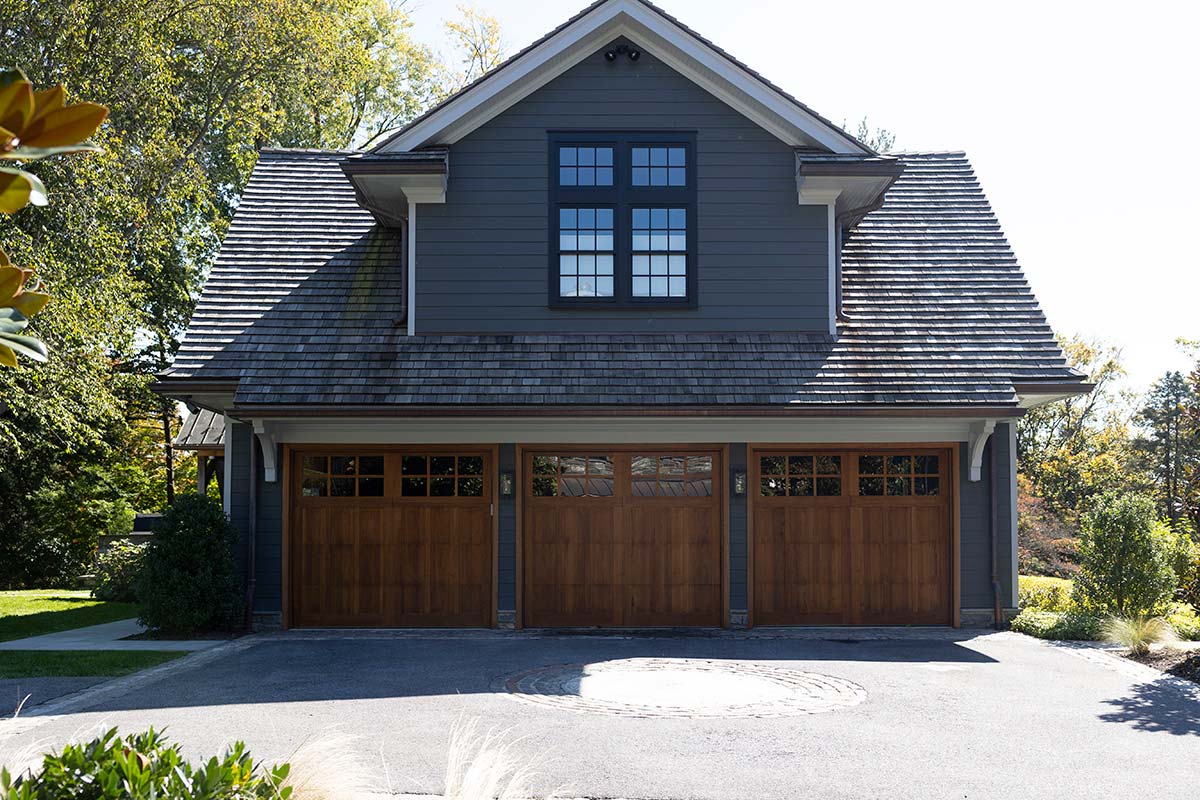 Modern garage with gray siding, shingled roof, and three wooden doors, surrounded by lush greenery and a paved driveway.