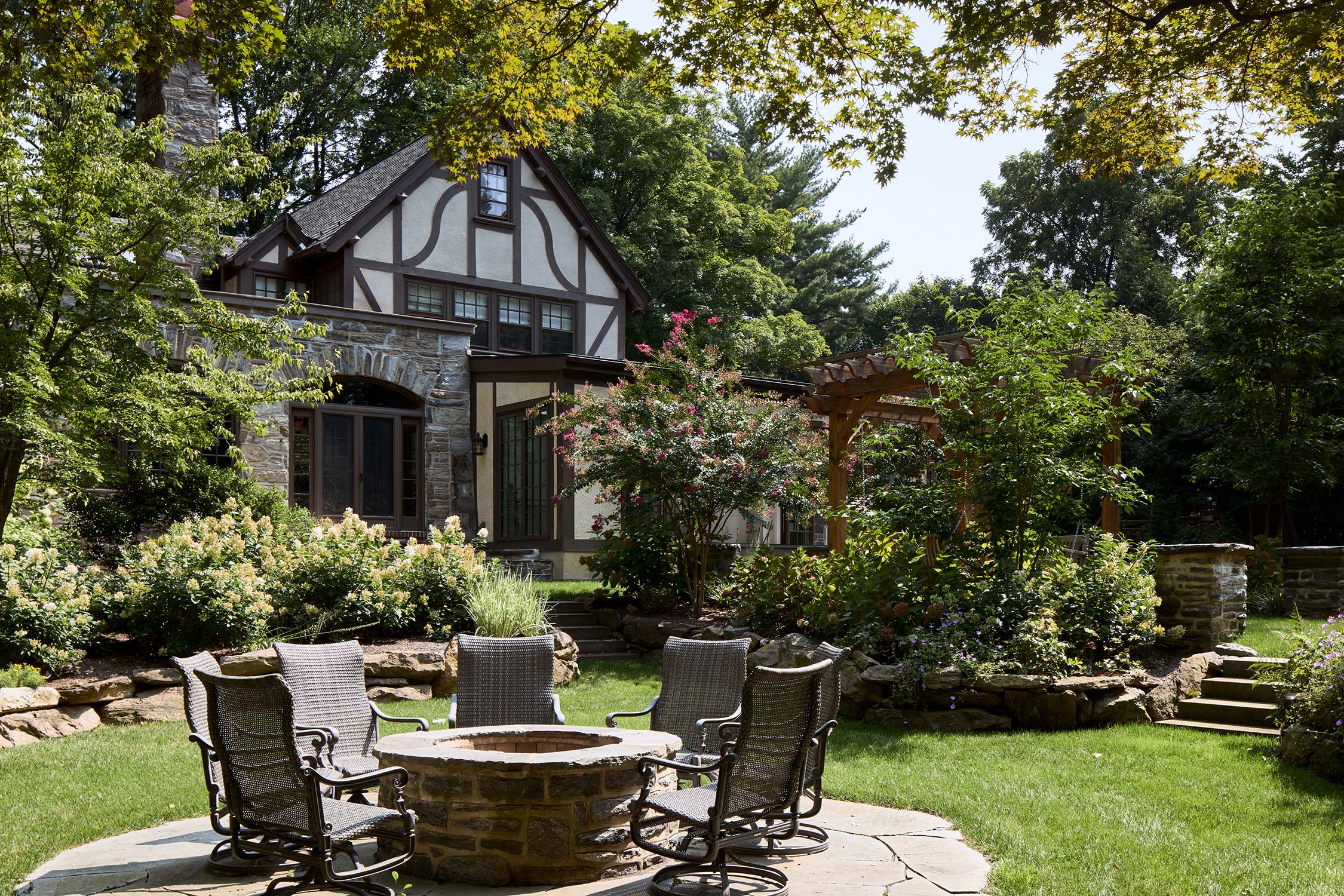 Stone house with Tudor-style gables, surrounded by a garden with a stone fire pit, wicker chairs, and wooden pergola.