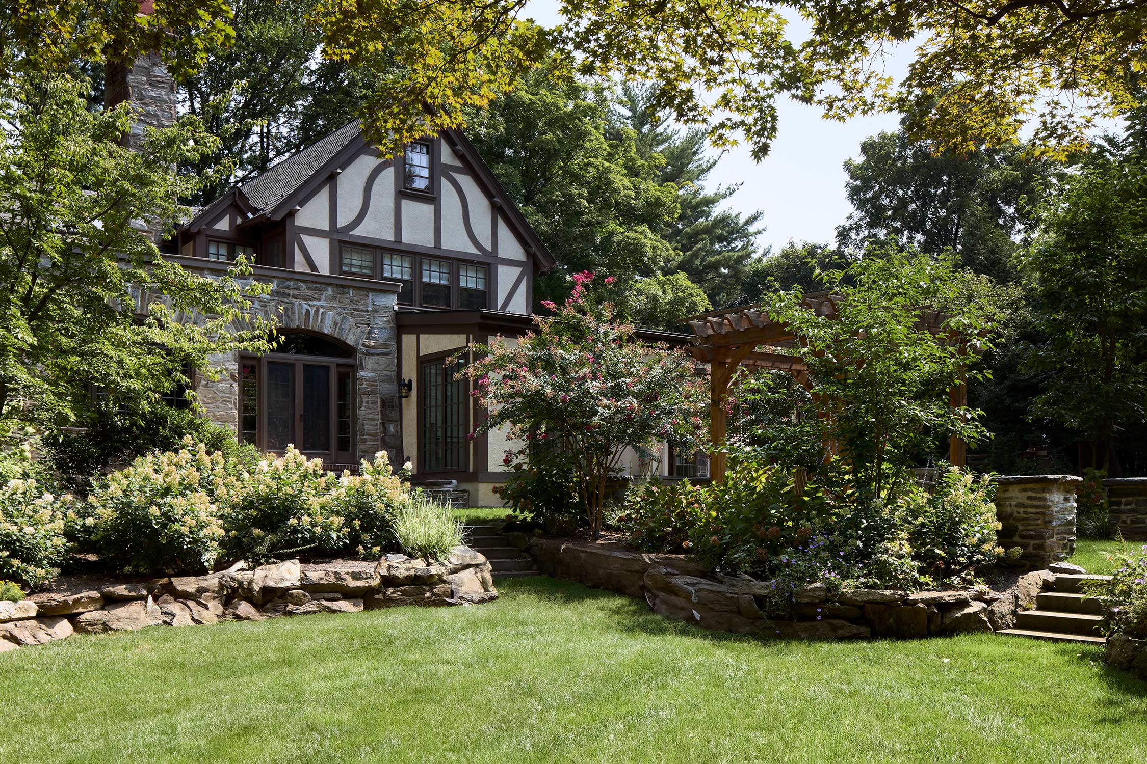 Charming Tudor-style home with stone and timber accents, surrounded by lush gardens, vibrant flowers, and a wooden pergola.