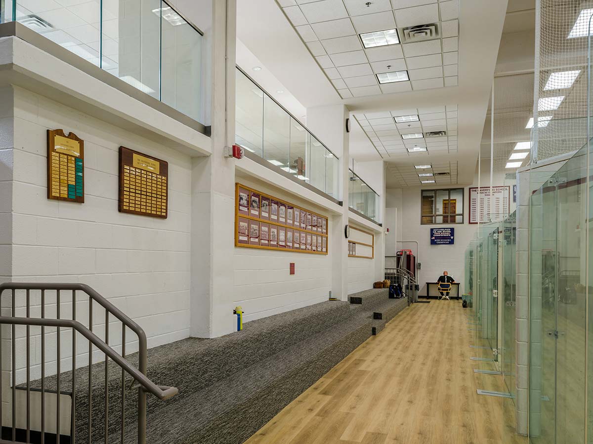 Indoor squash facility with glass-walled courts, bleachers, and a hardwood floor. Walls display championship banners and team photos from The Haverford School.