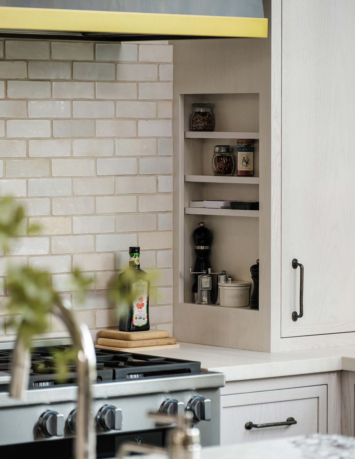 Kitchen scene with a gas stove, white brick backsplash, built-in shelves with spices, and a bottle on a wooden board.