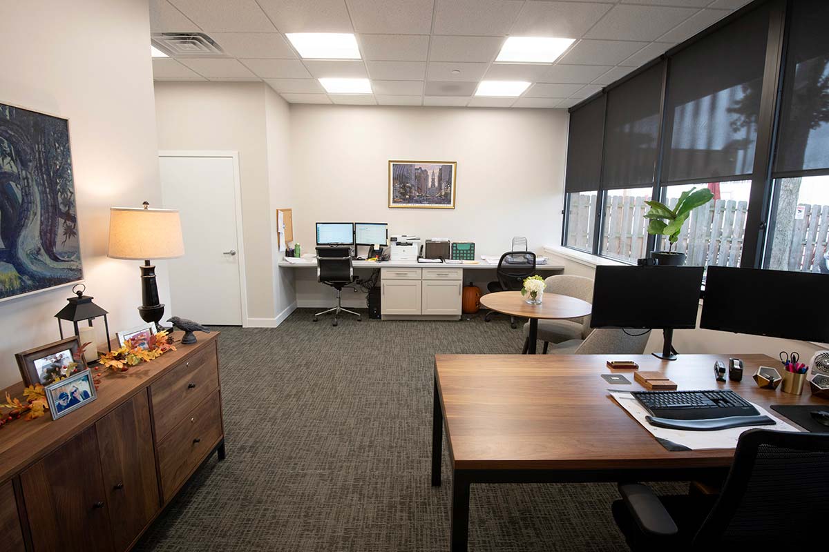 Office with wooden desk, dual monitors, chairs, framed photos, lamp, and plant by window.