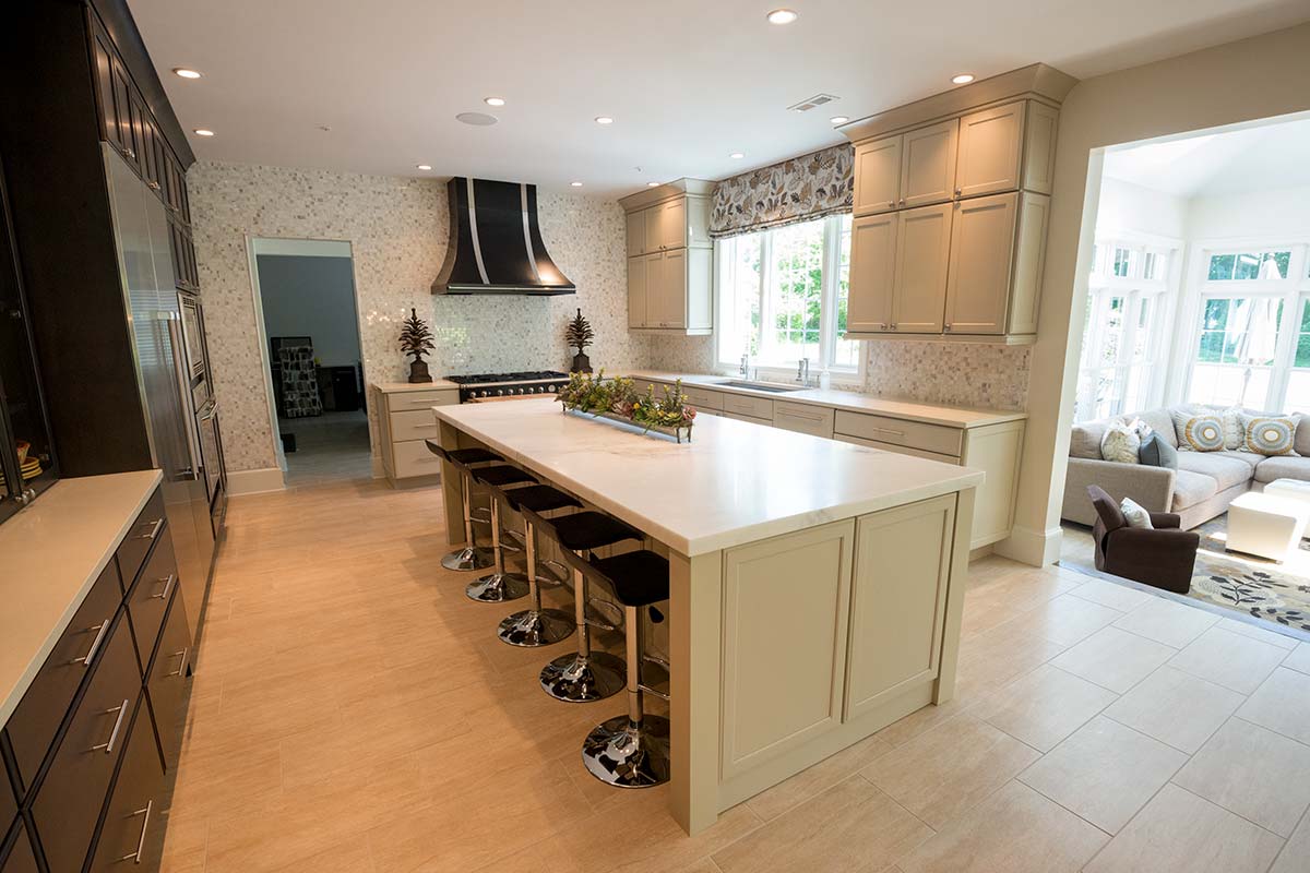 Bright kitchen with marble island, black range hood, light cabinets, and bar stools, opening to a cozy living area with large windows.