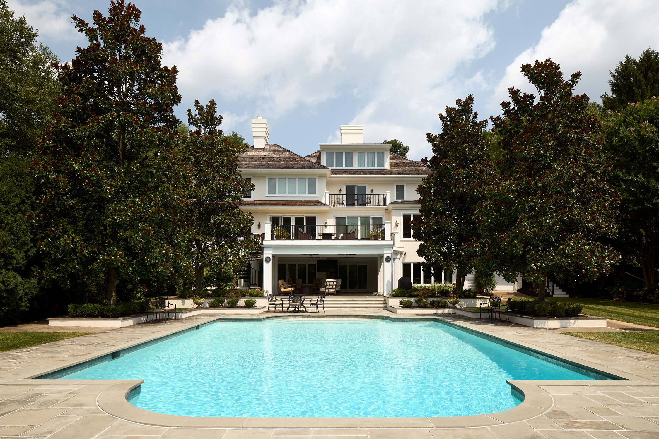 Large white house with pool, patio, and balcony, surrounded by trees and shrubs under a partly cloudy sky.