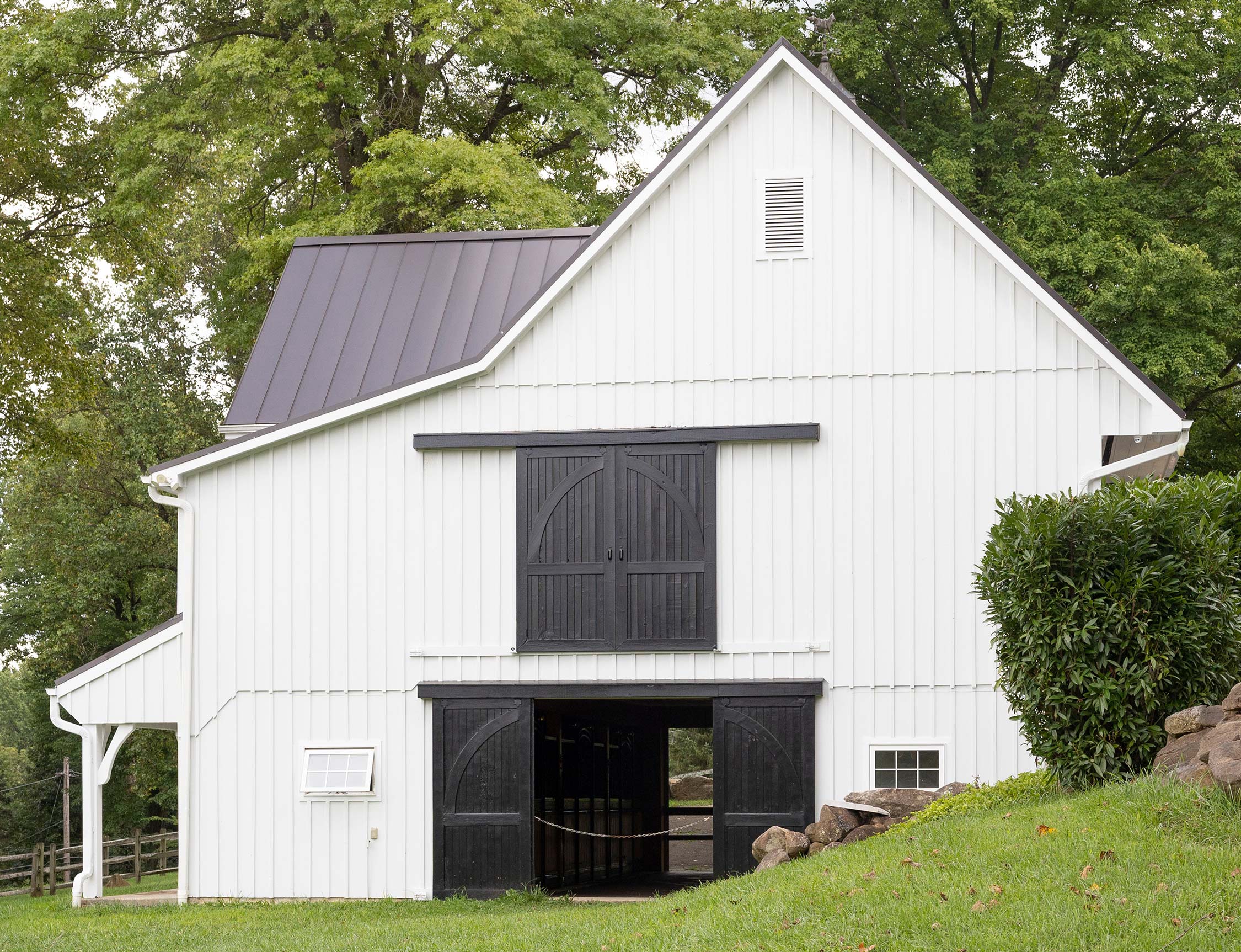 White barn with black double doors, metal roof, small windows, and surrounded by green lawn and trees on a hilly landscape.