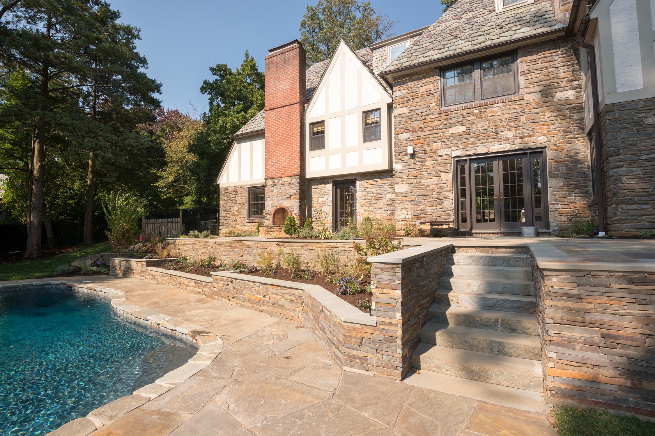 Stunning stone home with slate roof and brick chimney, overlooking a serene pool and terraced garden under a clear blue sky.