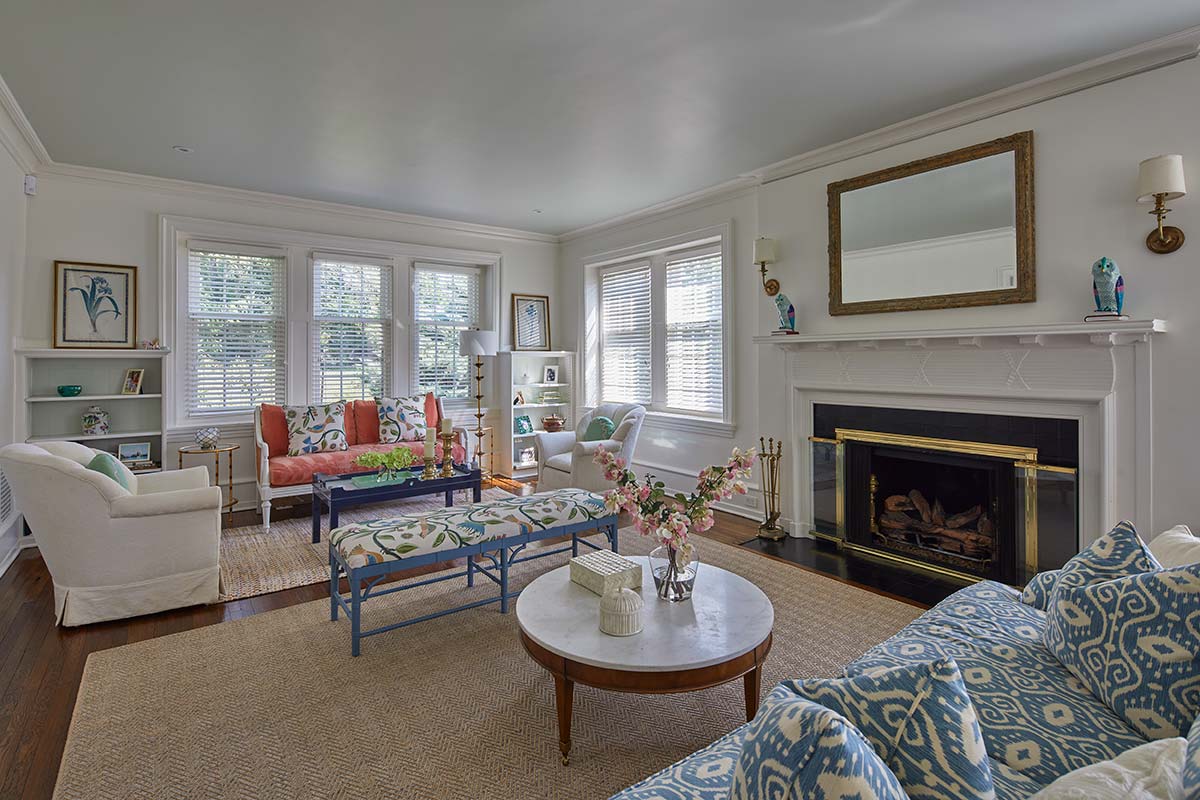 A bright living room with a fireplace, white walls, large windows, a coral sofa, blue ottoman, patterned blue sofa, and a marble coffee table with flowers.