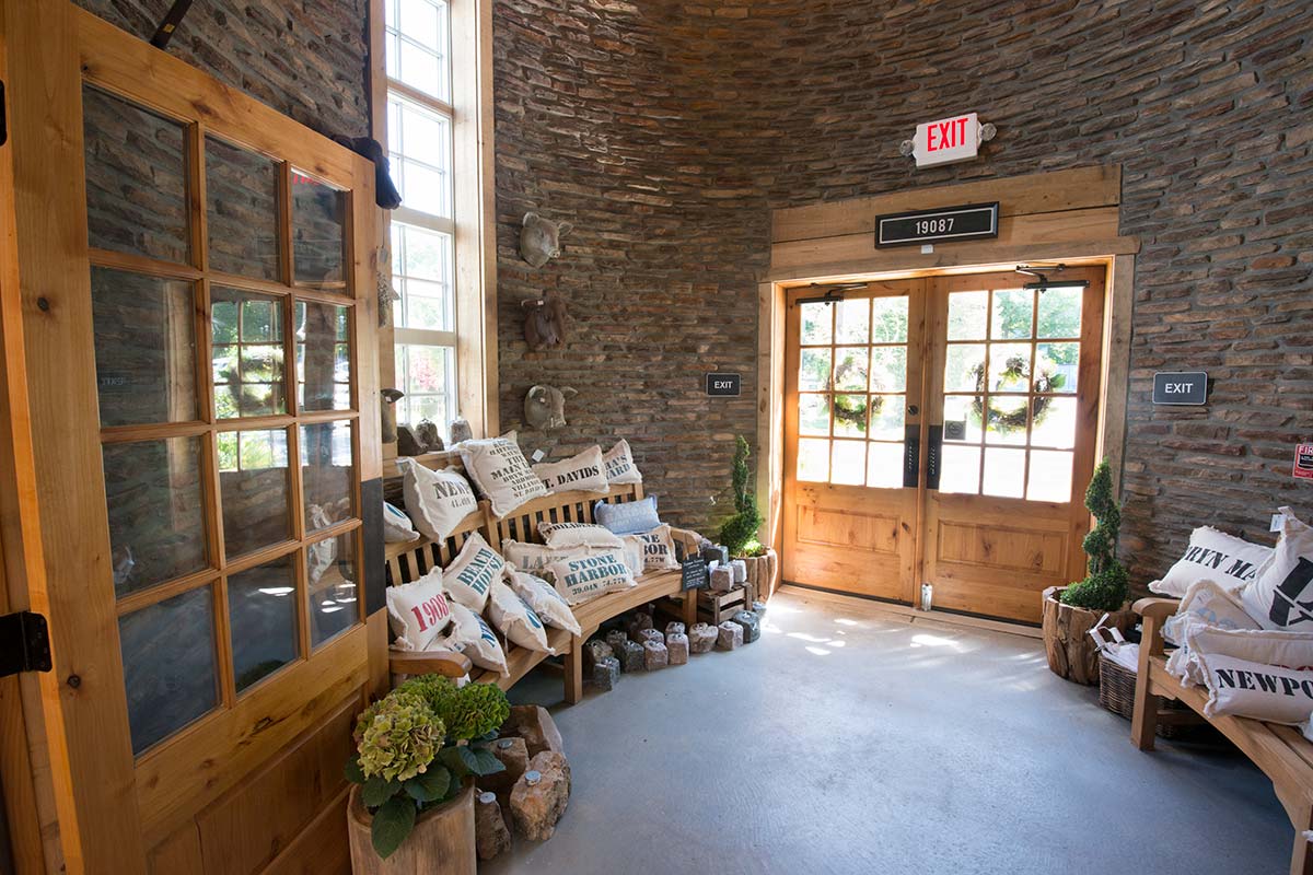 Stone-walled room with wooden doors, benches, decorative pillows, potted plants, and an exit sign above the door.