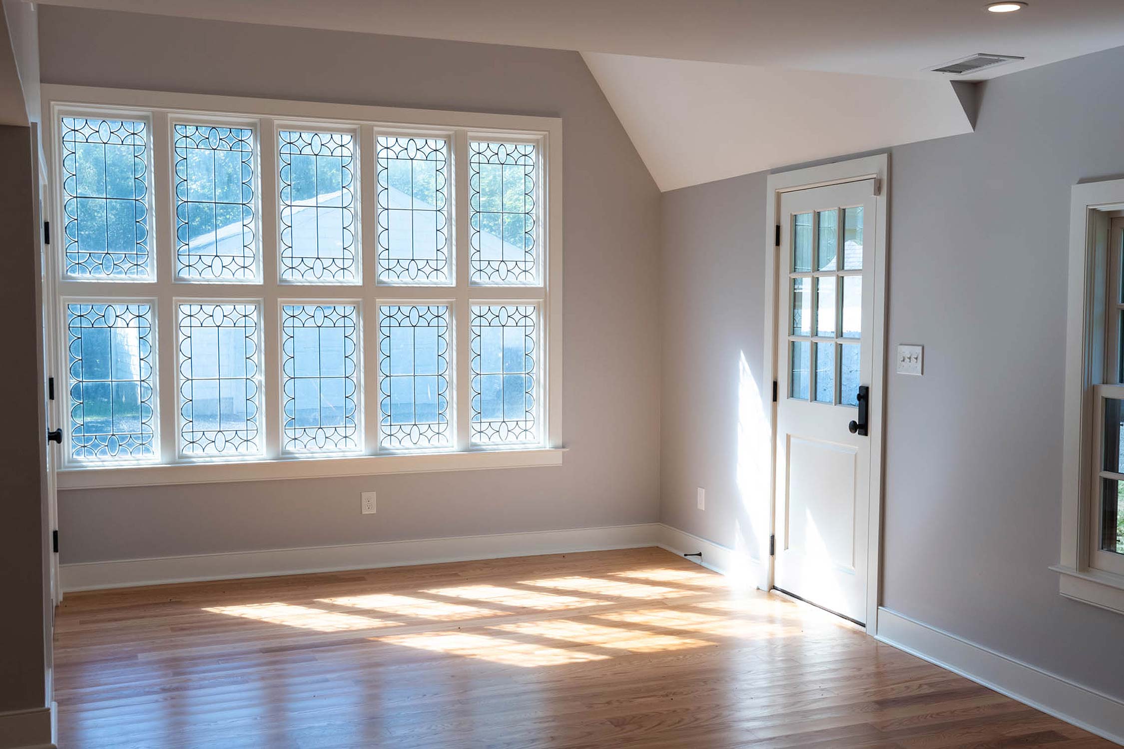 An empty room with hardwood floors, large decorative windows, a white door, and sunlight streaming in.