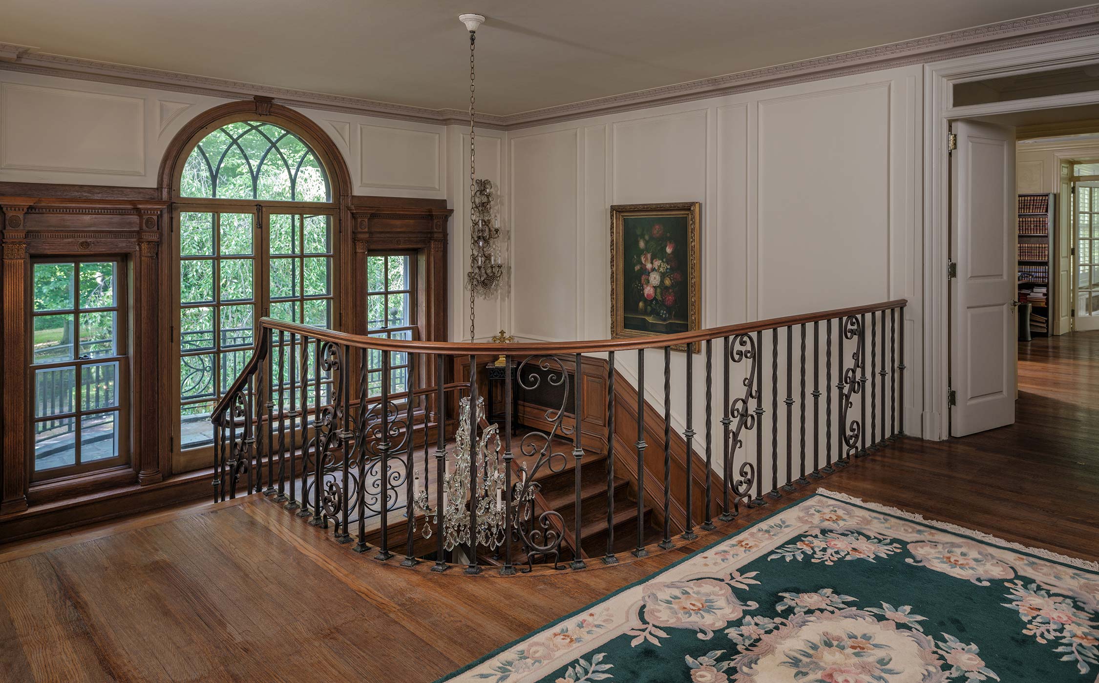Elegant landing with wooden staircase, wrought iron railing, and chandelier. Features large arched windows, a floral painting, and a patterned rug on hardwood floor.
