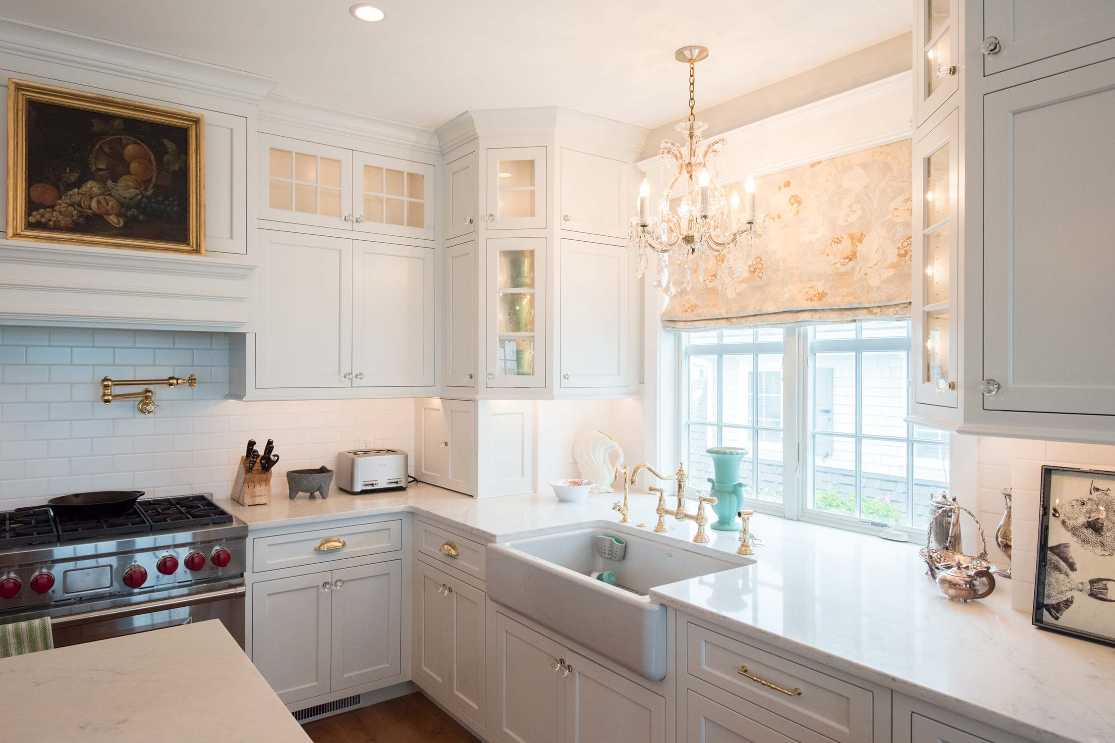 A bright kitchen with white cabinets, a chandelier, and a large sink by a window with floral curtains. A stove, toaster, and utensils are on the counter.