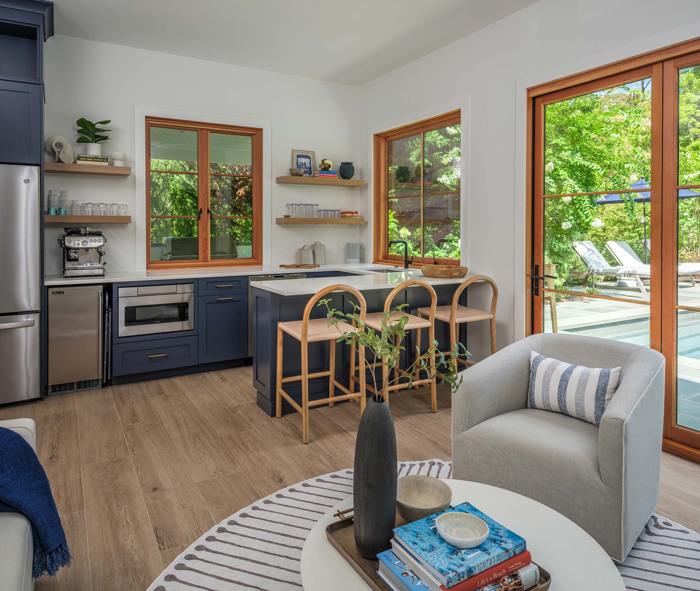 Modern kitchen with navy cabinets, white counters, bar stools, and a cozy living area with a gray chair, coffee table, books, and a vase. Large windows show a pool outside.