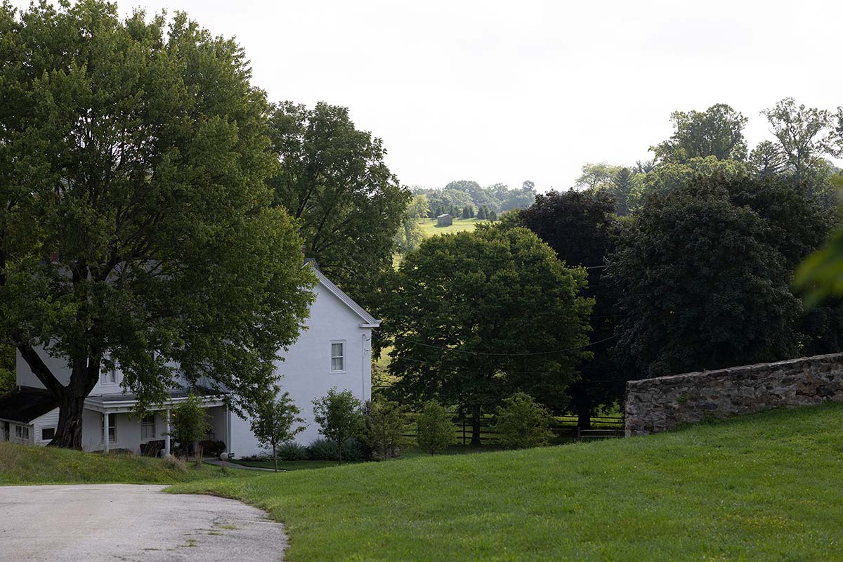 White house with porch and stone wall, surrounded by green lawn, trees, and fields under a light sky.