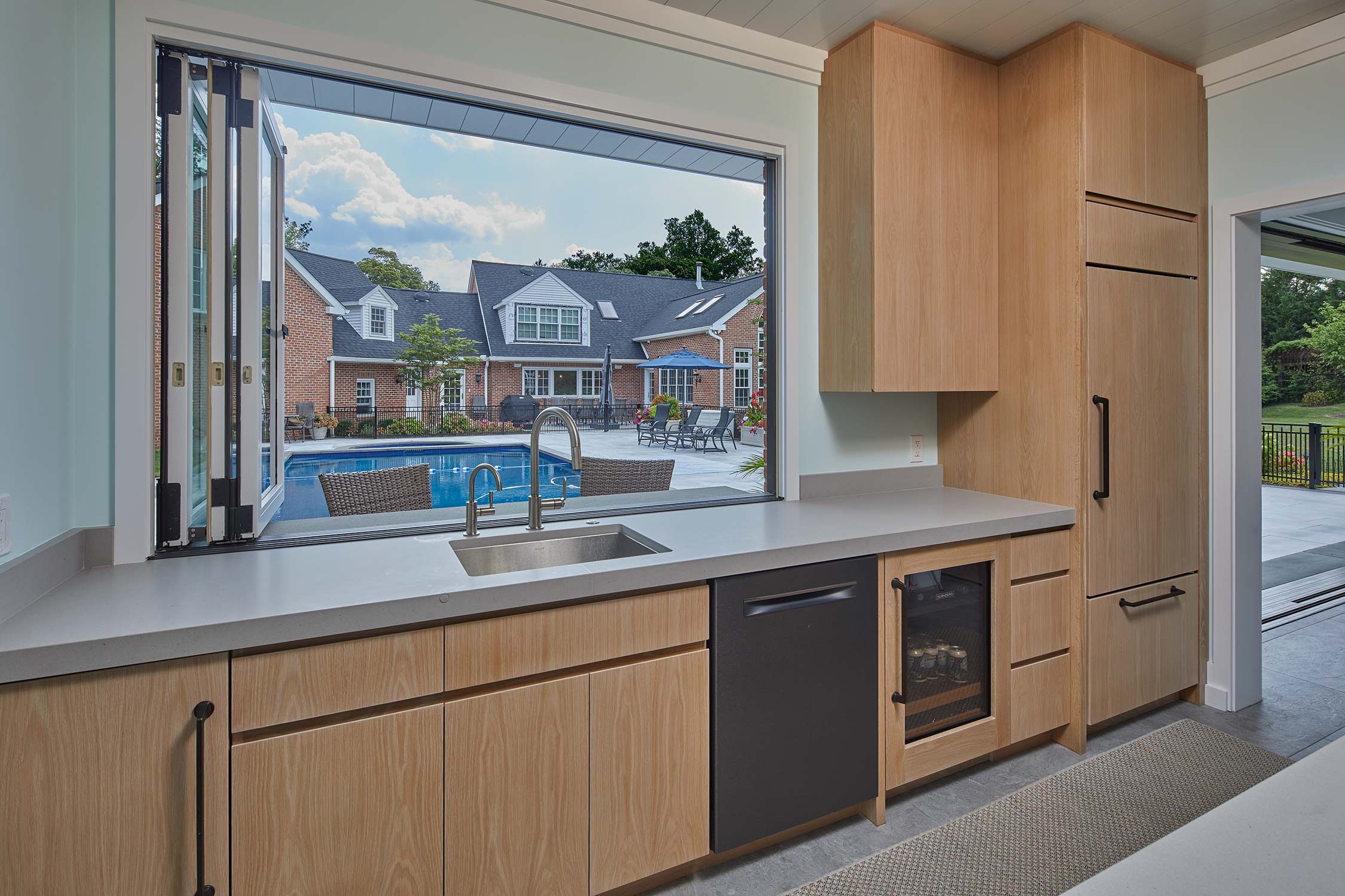 A modern pool house kitchen with wooden cabinets, a sink, and a dishwasher. Large windows open to a poolside patio with chairs and a brick house in the background.