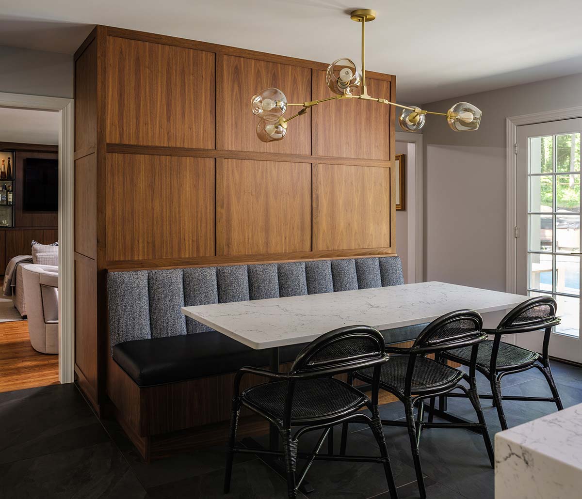 Dining area with a marble table, black wicker chairs, and a gray banquette against a wooden wall. A modern chandelier with glass bulbs hangs above.