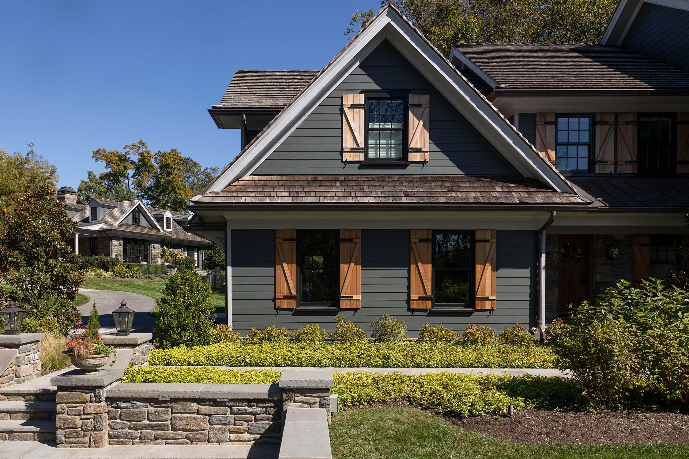 Modern house with gray siding, wooden shutters, and a shingled roof, surrounded by lush greenery, stone walls, and a well-maintained garden.