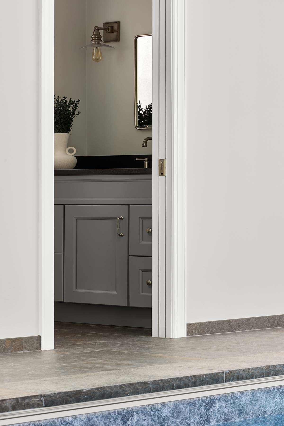 Bathroom next to a pool deck with gray cabinets, black countertop, white vase with greenery, mirror, and wall sconce.