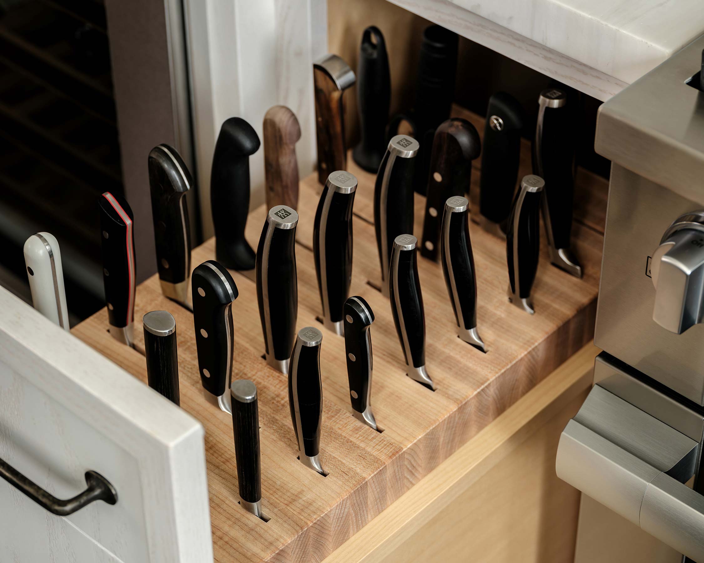 Wooden knife block with various knives stored in a kitchen drawer, next to a stainless steel oven and white cabinet.