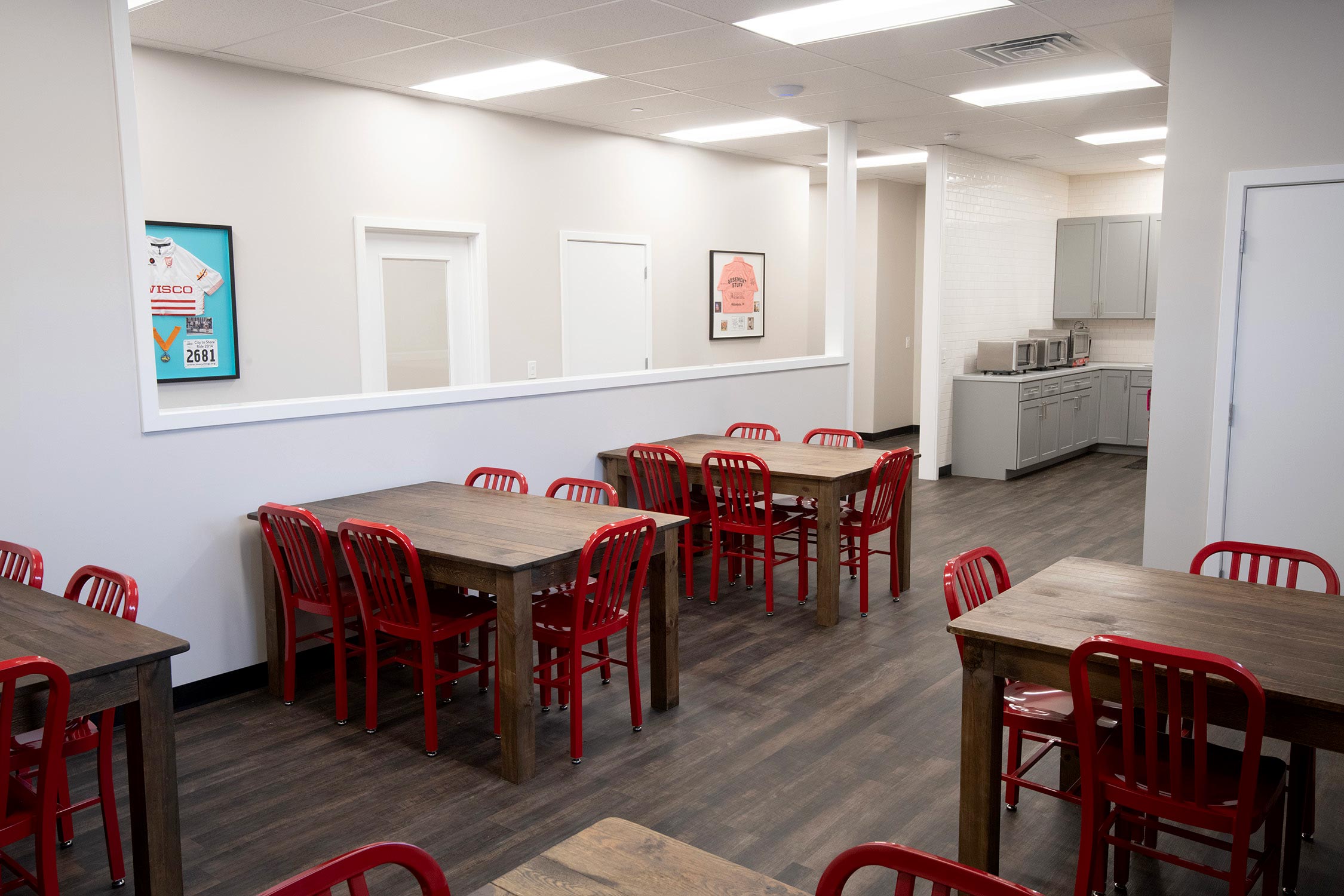 Break room with wooden tables, red chairs, kitchenette, and framed sports memorabilia on walls.
