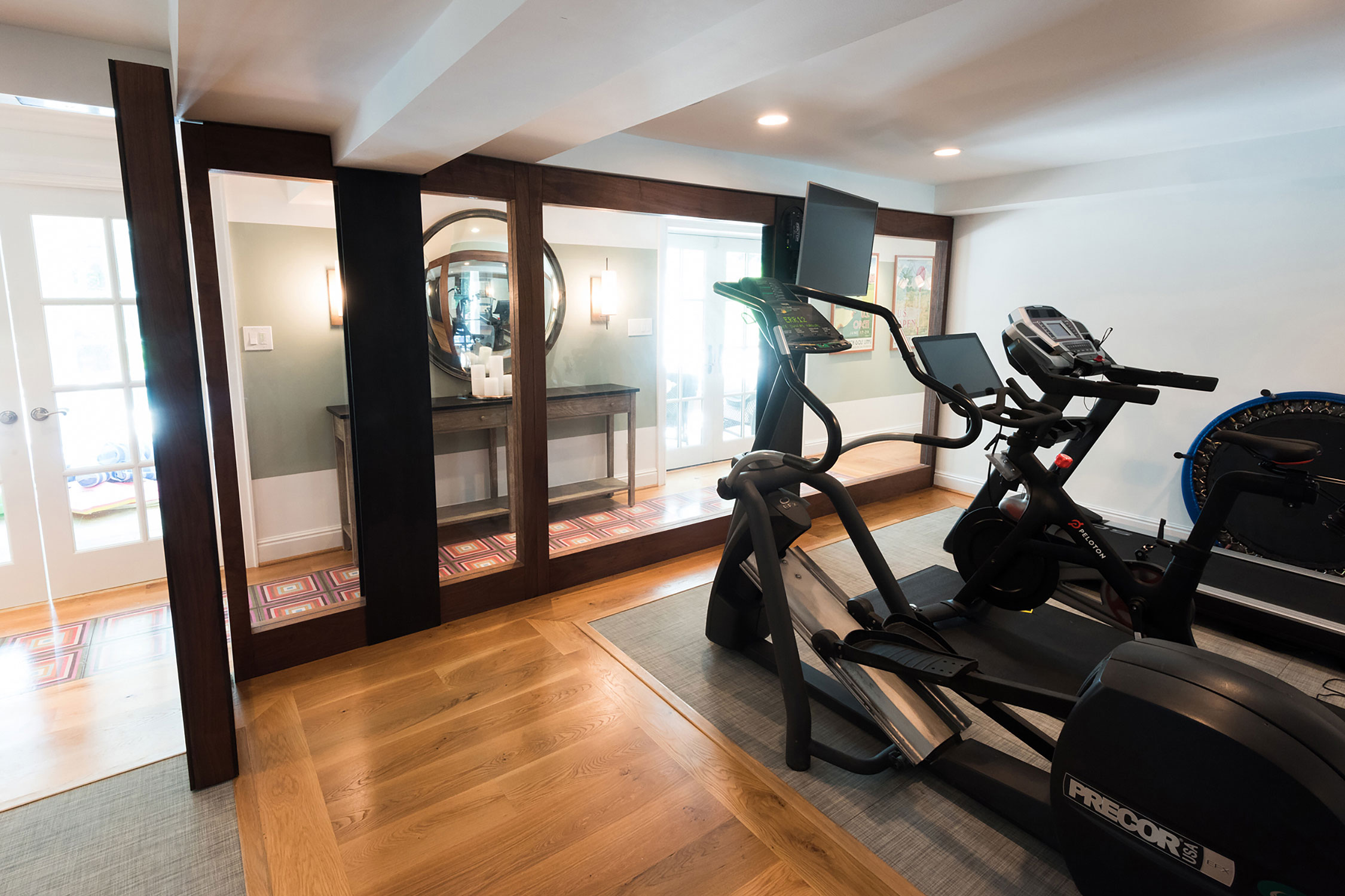 Modern home gym with a Precor elliptical, Peloton bike, and mirrored workout area. Features elegant wood flooring and a stylish console table.