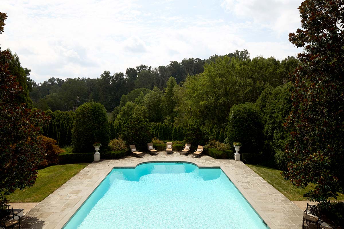 A backyard with a rectangular pool, stone patio, lounge chairs, decorative urns, and dense greenery.