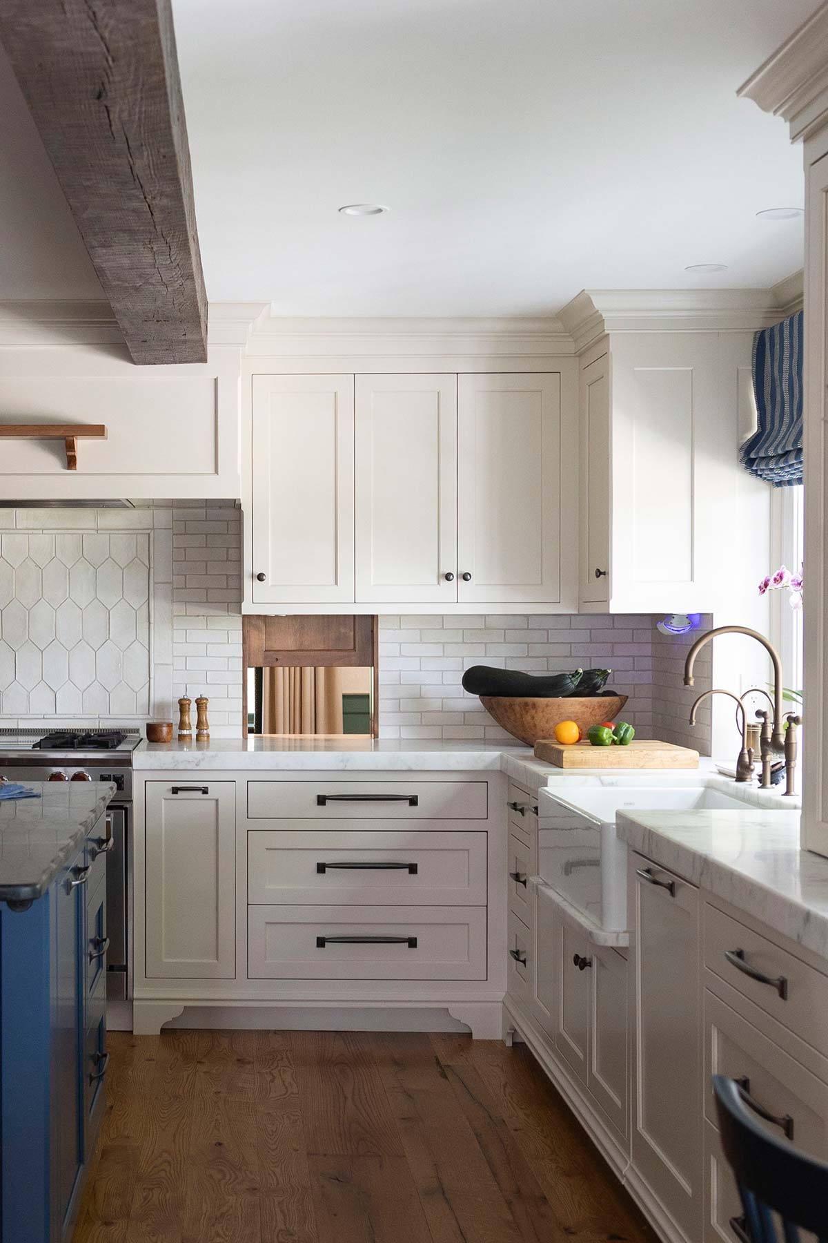 Modern kitchen with white cabinets, marble countertops, farmhouse sink, wooden beams, and a bowl of vegetables on the counter.
