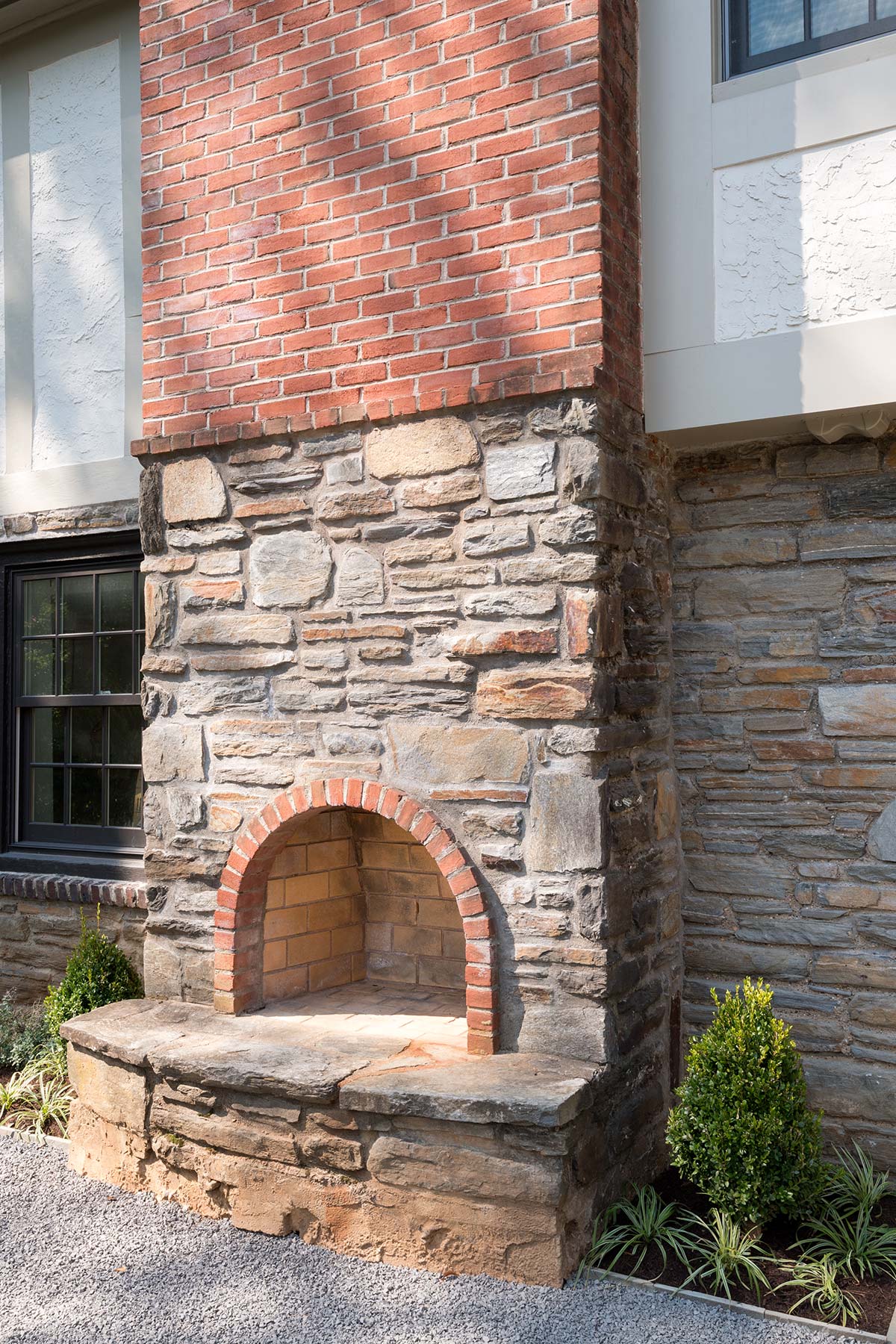 Rustic outdoor fireplace with brick and stone design, nestled in a landscaped garden with evergreen shrubs and gravel path.
