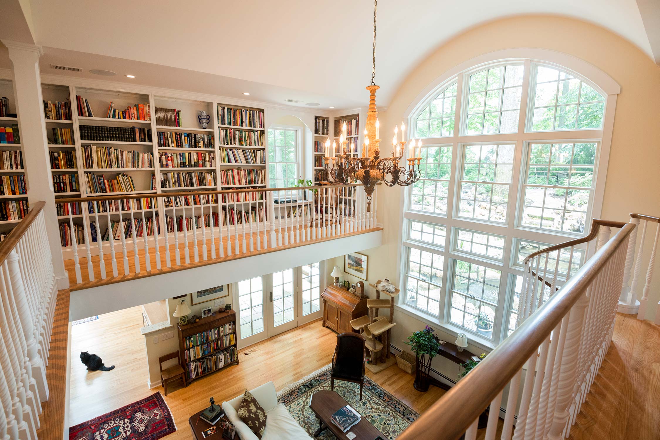Cozy library with high ceilings, a grand chandelier, and extensive bookshelves. Features large windows, elegant furniture, and a cat lounging. Ideal for reading.