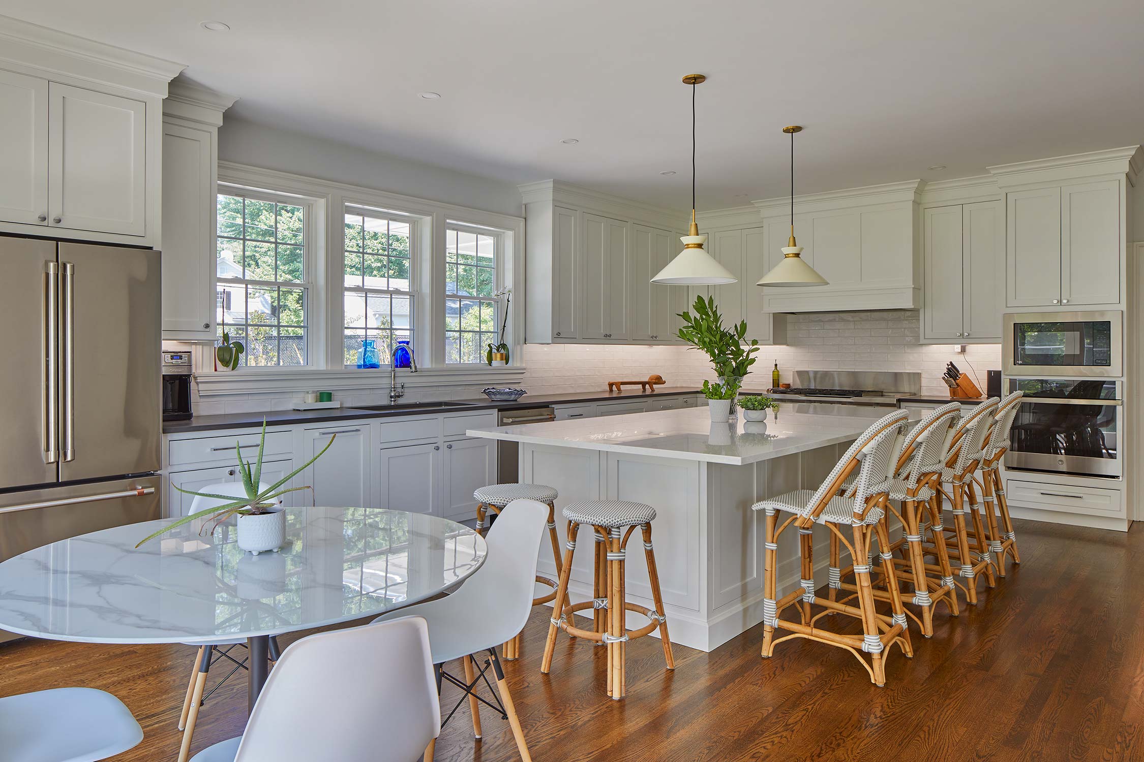 A modern kitchen with white cabinets, a marble island with wicker bar stools, pendant lights, stainless steel appliances, and a round dining table with a plant.